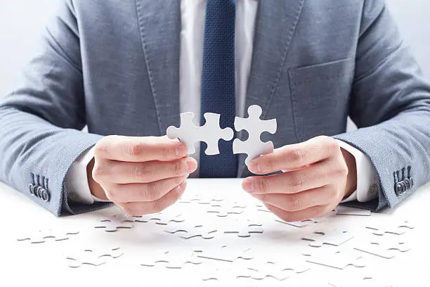 Businessman holding two pieces of puzzles with many other pieces on desk.